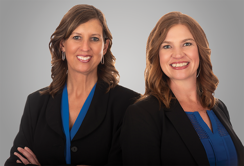 Two female professionals smiling at the viewer, wearing blue shirts, and black blazers.