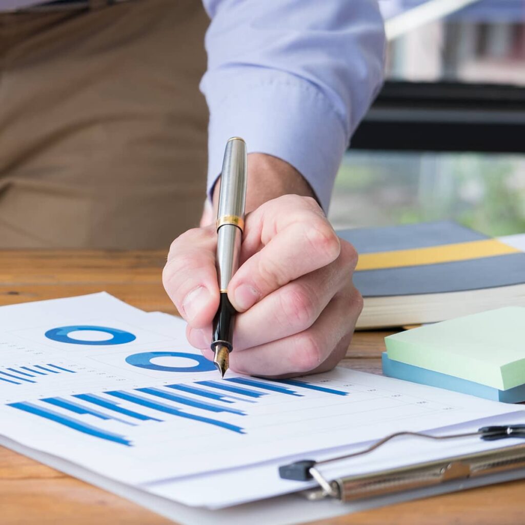 The hand of a male professional using a pen to make notes on a piece of paper with numerous charts and graphs on it.