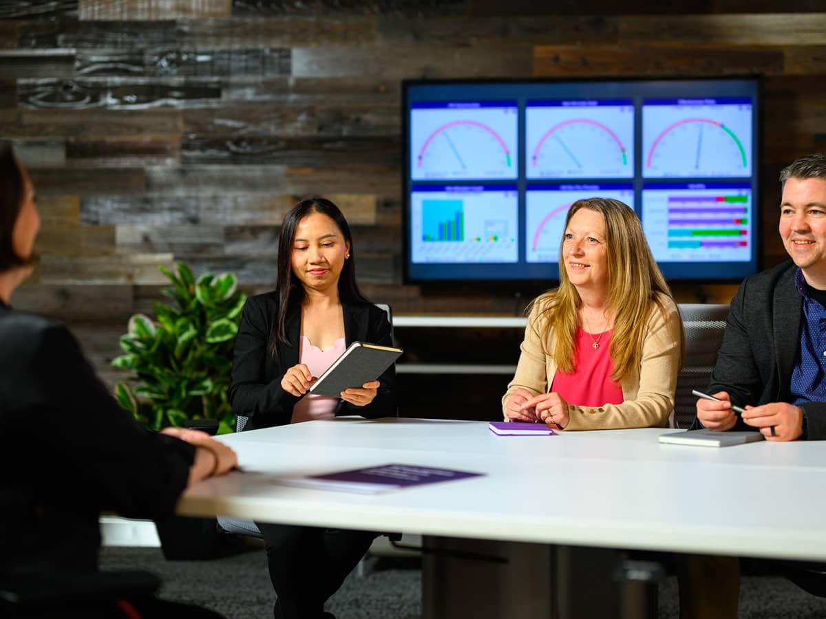 Three professionals meeting with a client in front of a television with graphs on it